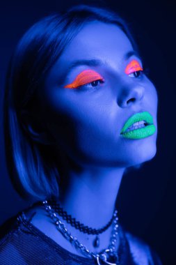 Renkli neon makyajlı ve kolyeli şık bir kadının koyu mavi arka planda başka yöne bakan portresi.