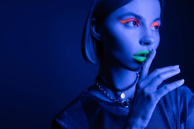 Neon makyajlı bir kadının portresi yeşil dudaklara dokunurken koyu mavi arka planda sessizlik işareti gösteriyor.