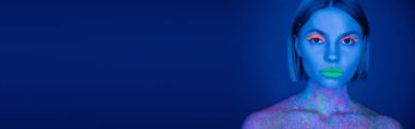portrait of woman with green neon lips and orange eye shadow looking at camera isolated on dark blue, banner clipart