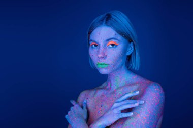 Canlı makyajlı, neon boyalı çıplak bir kadın koyu mavi renkli kameraya bakıyor.