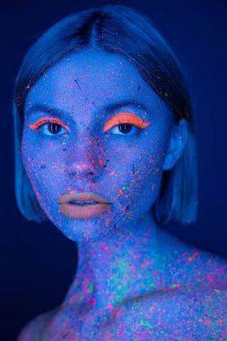 portrait of pretty woman with glowing makeup on face colored with neon paint looking at camera isolated on dark blue clipart
