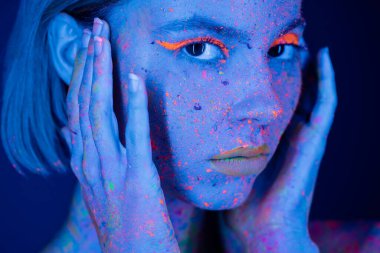 portrait of woman in neon makeup and fluorescent paint holding hands near face on dark blue  clipart