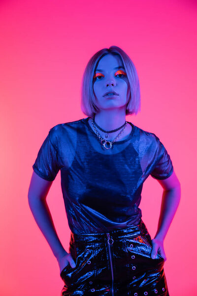 fashionable woman standing with hands in pockets of leather skirt on deep pink background with blue neon light
