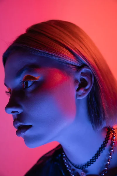 portrait of young woman with necklaces and neon makeup in blue light on pink and coral background