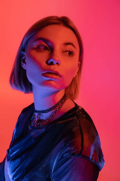 portrait of woman in necklaces looking away in neon light on pink and coral background