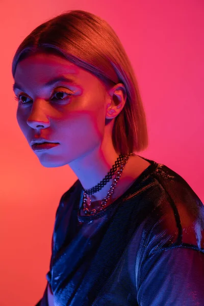 portrait of trendy woman in necklaces posing in neon light on coral and pink background