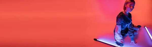 stock image trendy woman sitting near purple fluorescent lamps on coral background, banner