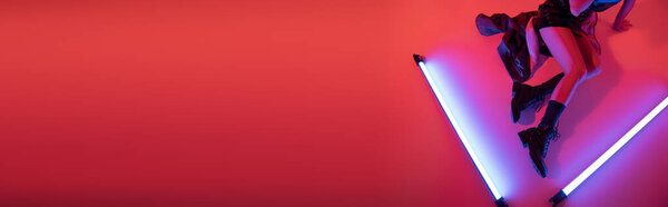 top view of cropped woman in boots sitting near purple neon lamps on carmine red background, banner
