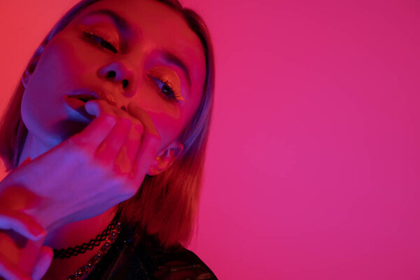 portrait of young woman with neon makeup holding hand near face on deep pink background