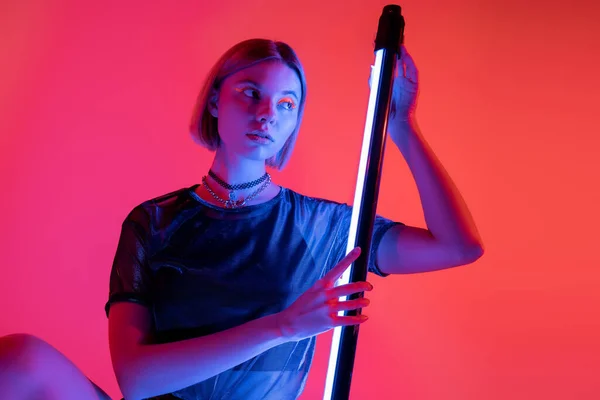 stock image young and trendy woman posing in blue light of neon lamp on coral and pink background