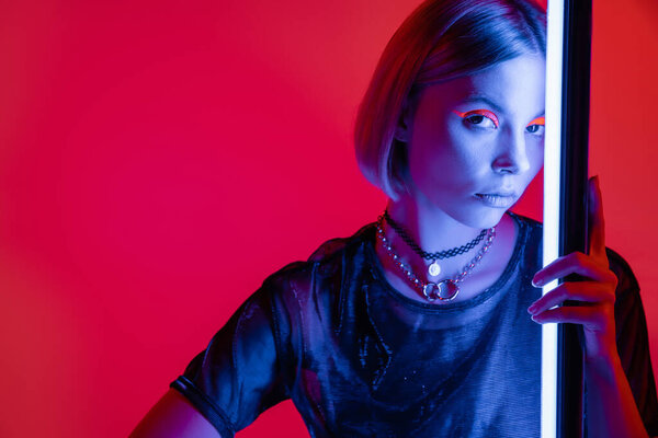 stylish woman in bright makeup and necklaces looking at camera in blue light of neon lamp on carmine red background