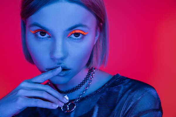 sensual woman with glowing makeup touching lips in blue neon light on coral pink background