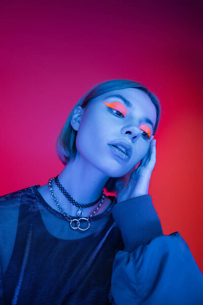 trendy woman in necklaces and neon makeup holding hand near face on pink and red background