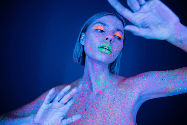 nude and sensual woman with neon makeup and colored body posing on dark blue background