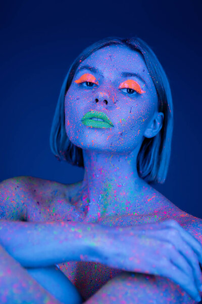 portrait of young woman in bright neon makeup and colorful body paint looking at camera isolated on dark blue