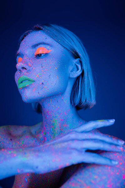 young woman with neon makeup touching body colored in fluorescent paint isolated on dark blue