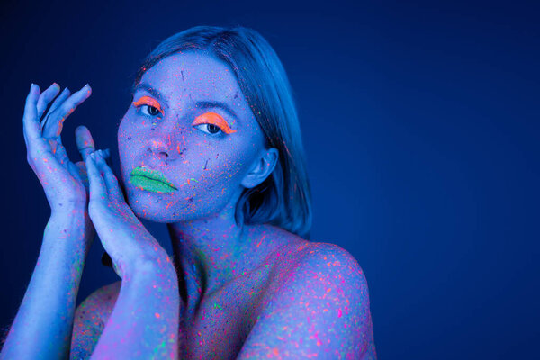woman in vibrant makeup and colorful neon splashes on body looking at camera isolated on dark blue