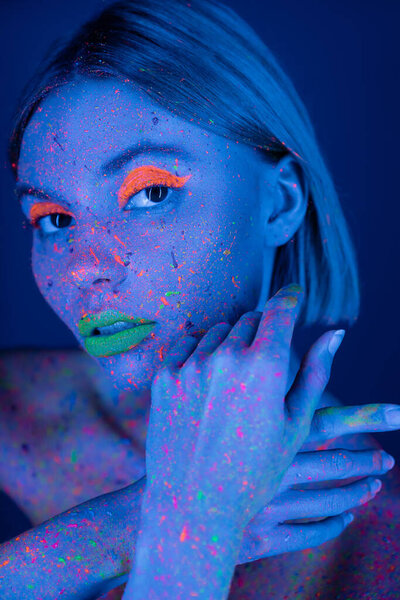 woman with glowing makeup and paint splashes posing in neon light isolated on dark blue