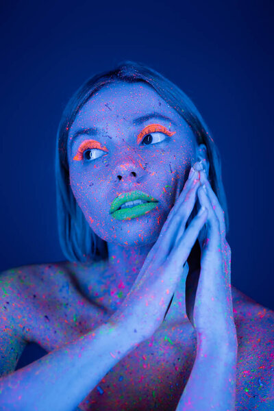 young woman with vibrant neon makeup and bright paint splashes on body looking away isolated on dark blue