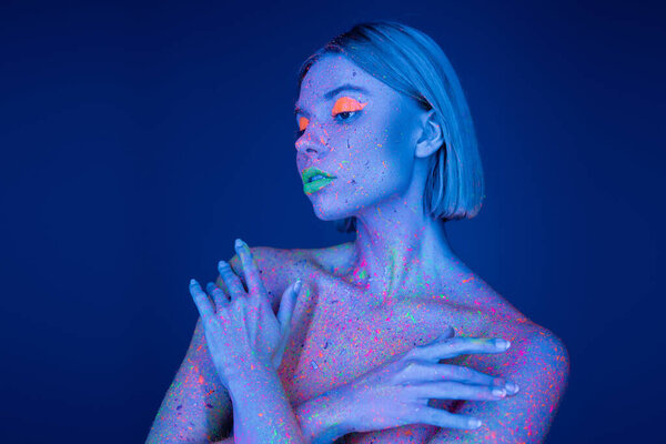 nude woman in neon makeup and glowing paint splashes on body posing with crossed arms isolated on blue