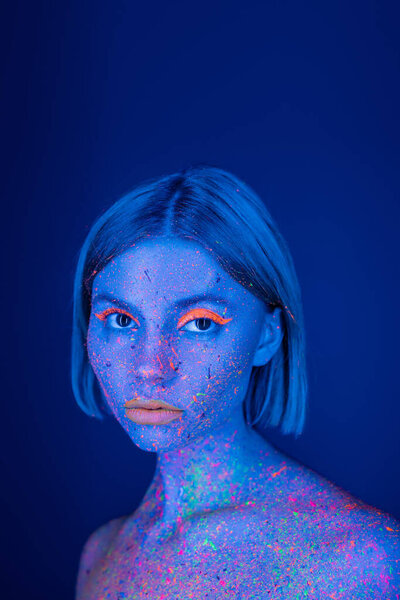 portrait of woman with glowing makeup and neon paint splatters on body isolated on dark blue