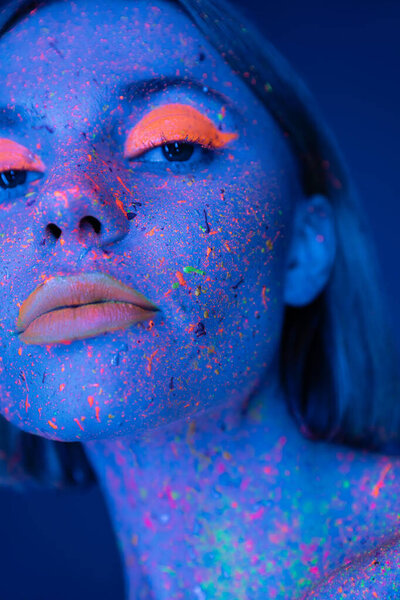 close up portrait of woman in glowing neon makeup and bright paint isolated on dark blue