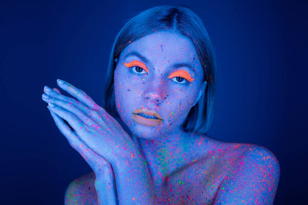 pretty woman in neon makeup and colorful paint looking at camera isolated on dark blue