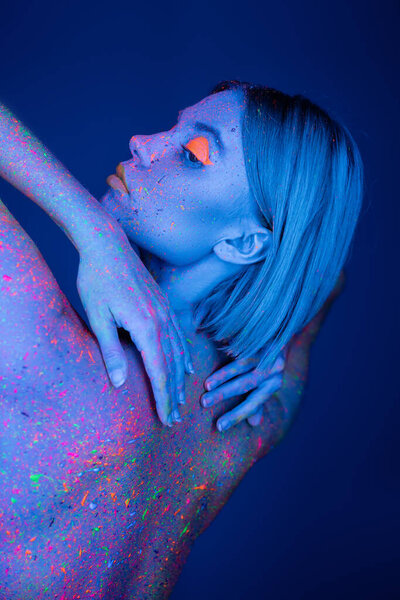 sensual woman in colorful neon body paint touching back isolated on dark blue