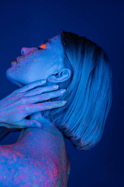 side view of woman in glowing neon body paint touching neck isolated on dark blue