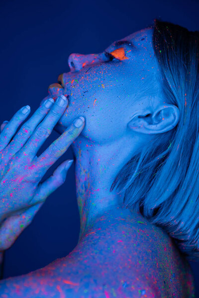 side view of woman in neon makeup and body paint holding hands near face isolated on dark blue