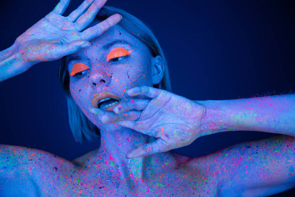 young woman in glowing makeup and neon body paint posing with hands near face isolated on dark blue