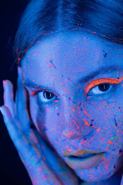 close up portrait of woman with neon makeup and colorful paint on face looking at camera isolated on dark blue