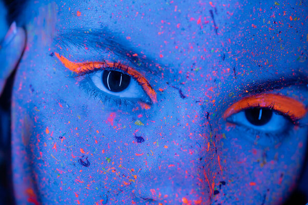 close up view of cropped woman with neon eye shadow and vibrant paint splatters looking at camera in blue light
