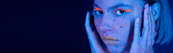 portrait of woman in vibrant makeup and neon splashes holding hands near face isolated on dark blue, banner