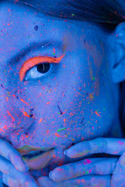 partial view of woman holding hands near face with neon makeup and bright paint splashes in blue light 
