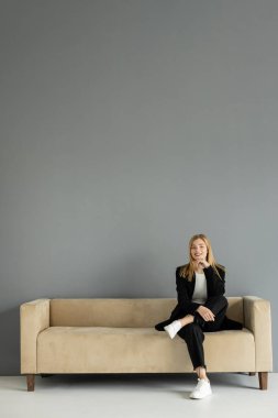 Cheerful blonde woman looking at camera while sitting on couch near grey wall  clipart