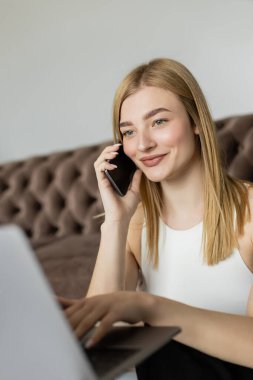 Gülümseyen sarışın koçun portresi cep telefonuyla konuşuyor ve oturma odasında bulanık dizüstü bilgisayar kullanıyor. 