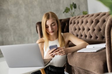 Güzel sarışın koç dizüstü bilgisayarın yanında akıllı telefon ve kanepede defter kullanıyor. 