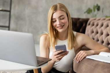 Genç koç oturma odasında bulanık dizüstü bilgisayar ve defter yanında cep telefonu kullanıyor. 