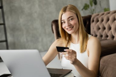 Blonde woman smiling at camera while holding credit card near laptop in living room  clipart