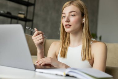 Young coach in wireless earphone holding pen during video chat on laptop near blurred notebook at home  clipart