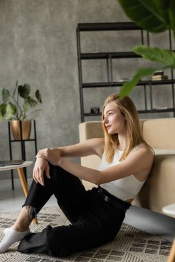 pensive blonde woman in black jeans sitting on floor near sofa and looking away in loft style apartment clipart