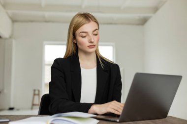 Siyah ceketli genç sarışın koç laptopta çalışıyor. Bulanık defterin yanında.