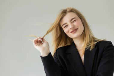 Siyah ceketli neşeli sarışın kadın saçlarını çekiyor ve gri renkli kameraya göz kırpıyor.