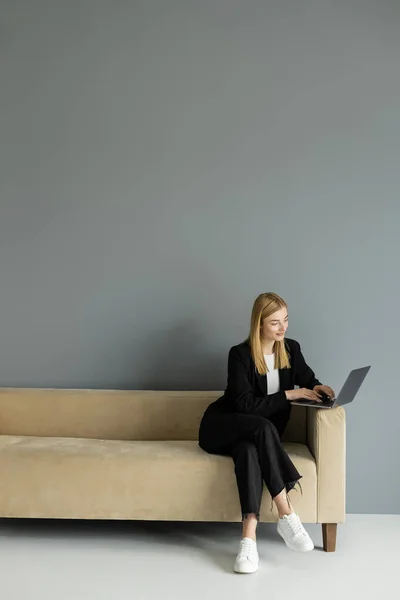 stock image Smiling blonde coach using laptop while sitting on beige sofa at home 