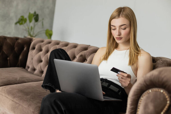Blonde woman in top holding credit card and using laptop on comfortable couch 