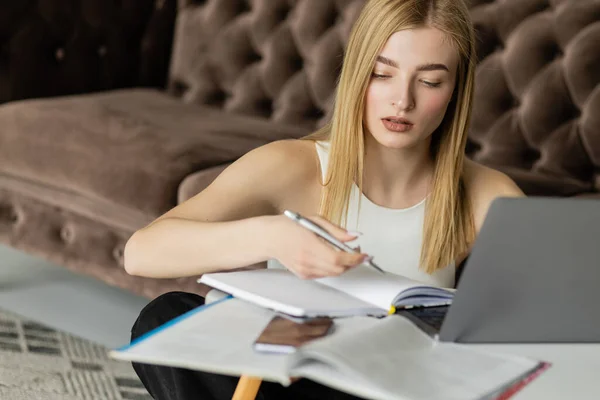 Młoda Kobieta Patrząc Notatnik Pobliżu Urządzeń Podczas Edukacji Online Salonie — Zdjęcie stockowe
