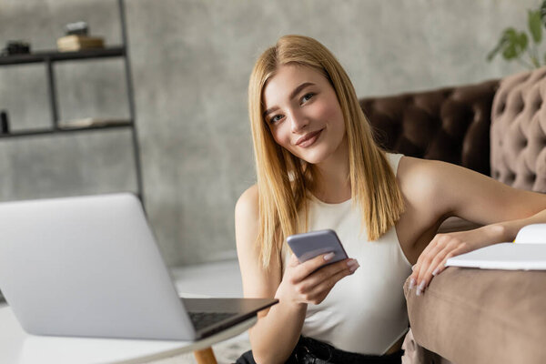 Блондинка тренер держит смартфон и смотрит на камеру возле ноутбука и ноутбука дома 