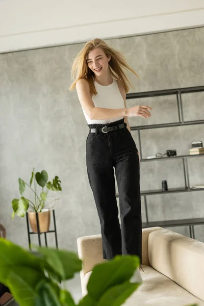 young excited woman in black jeans dancing on couch in modern apartment