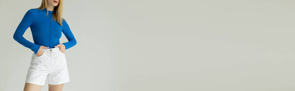 partial view of woman in blue long sleeve shirt standing with hands in pockets of white shorts isolated on grey, banner